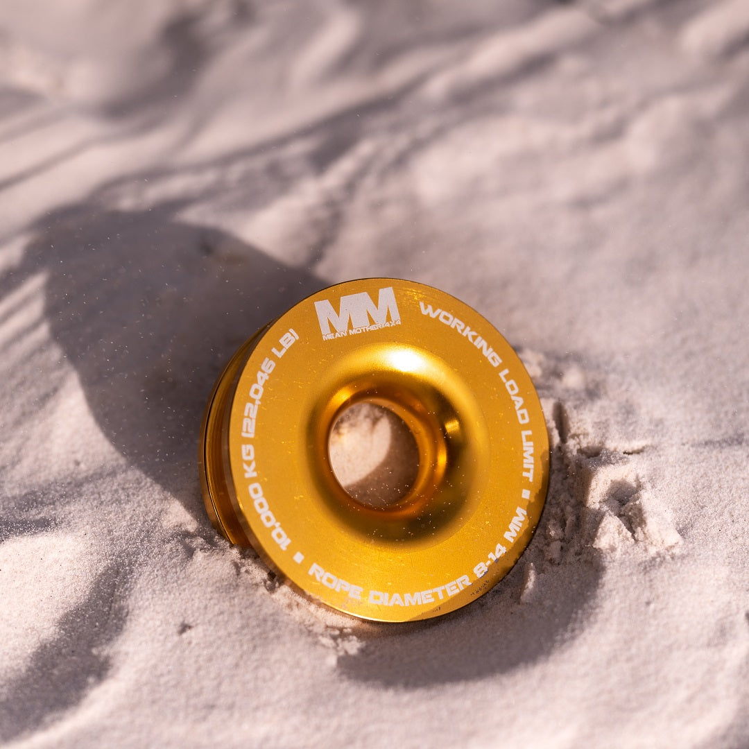 A partially buried Snatch Ring 10,000kg by Mean Mother 4x4 gleams under the sun on sand. Its engraved specs include "Working Load Limit" and "Rope Diameter 9.4 mm," along with the "MM" logo, highlighting its compatibility with synthetic winch ropes as a recovery accessory.