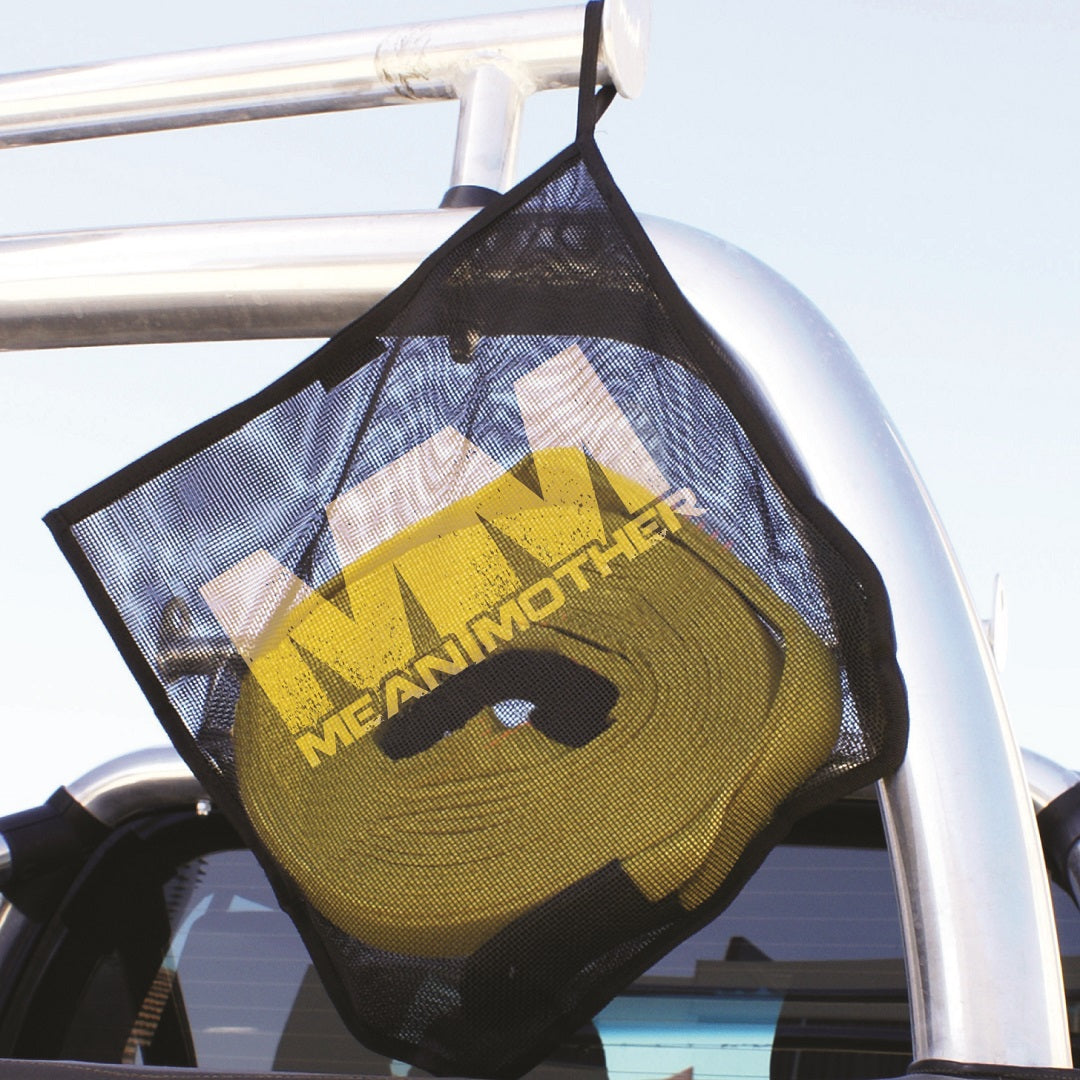 A Mean Mother Snatch Strap 11,000kg is stored inside a black mesh drying bag, suspended from a metal bar on the back of a vehicle. The strap is yellow and features the distinctive "MEAN MOTHER" branding. The setup includes reinforced eyelets and durable nylon webbing, set against the backdrop of a clear blue sky.