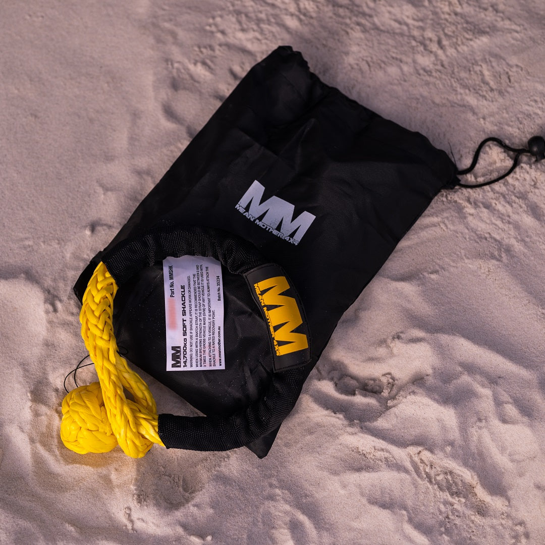 A black drawstring bag labeled "Mean Mother 4x4" rests on the sand, containing a vibrant yellow UHMPE synthetic rope peeking out. Shadows cast by the sunlight reveal the grainy texture of the sand, hinting at its role in a versatile recovery kit featuring the Mean Mother Soft Shackle 14,700kg Twin Pack.