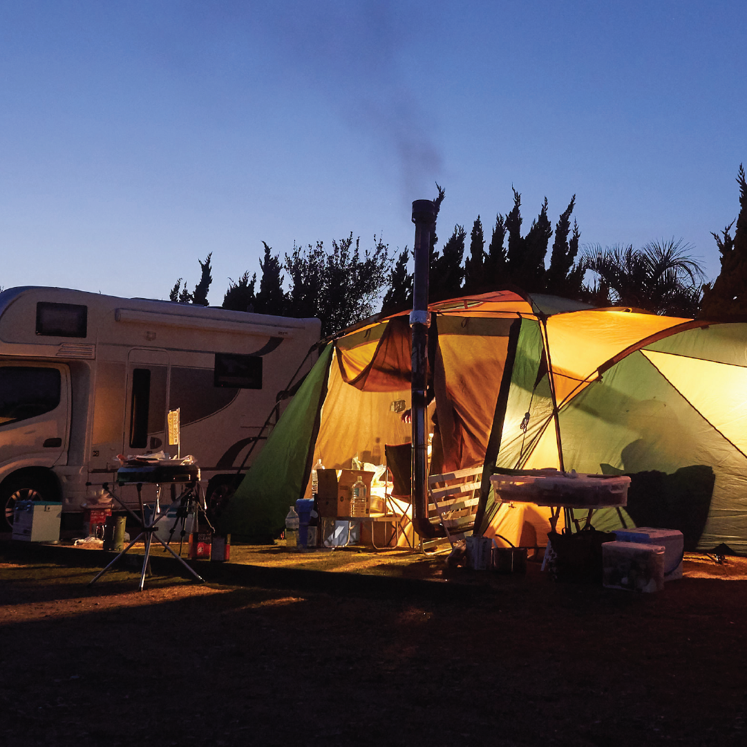 At dusk, a glowing tent and nearby camper van create a picturesque campsite. The serene atmosphere is enhanced by the MEAN MOTHER Led Light Bar Kit - Single Colour 12V from Mean Mother 4x4, illuminating the scene. Cooking supplies and chairs are arranged in front of the tent, while trees form striking silhouettes against the evening sky.