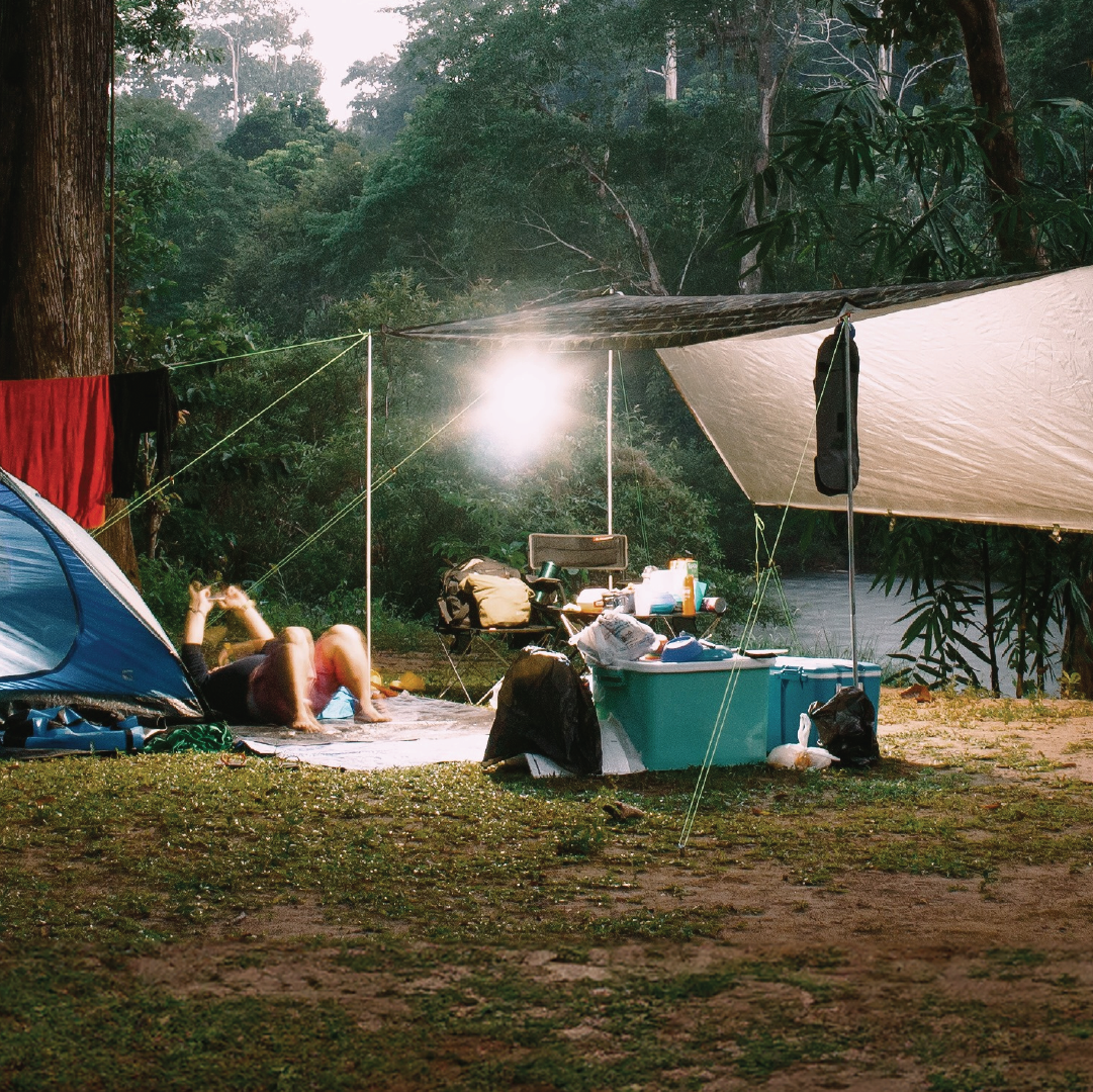 A campsite showcases a tent and tarp shelter, with someone lounging on the ground, either reading or using a device. Clothes are hung out to dry on a line, while various gear is strewn about. In the background, trees and a river provide a picturesque setting, illuminated by the gentle glow of waterproof MEAN MOTHER Led Light Bar Kit - Single Colour 12V from Mean Mother 4x4.