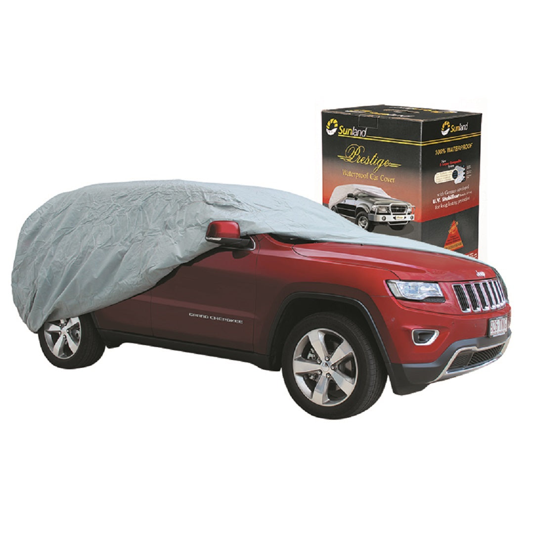 A red SUV is partially wrapped with a silver Prestige waterproof fabric car cover. In the background, a box labeled "Prestige" features an image of a fully covered 4WD car with product details, showcasing UV-stabilized protection.