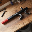 A Sperling Steering Wheel Lock Prolock with a red handle and grey adjustable grips, resembling an anti-theft device, rests on a cluttered wooden workbench with tools and paint cans in the background.