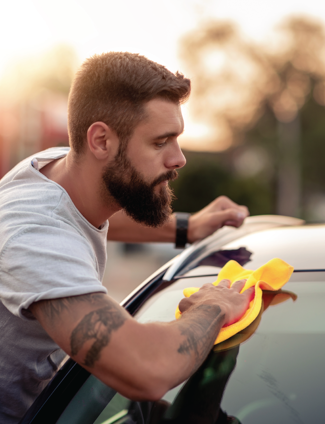 At sunset, a bearded man in a gray t-shirt with arm tattoos uses the Sunland-Protection CHAMOIS SYNTHETIC PVA (43 x 32cm), diligently cleaning a car windshield. He focuses on achieving a streak-free finish against the blurred background.