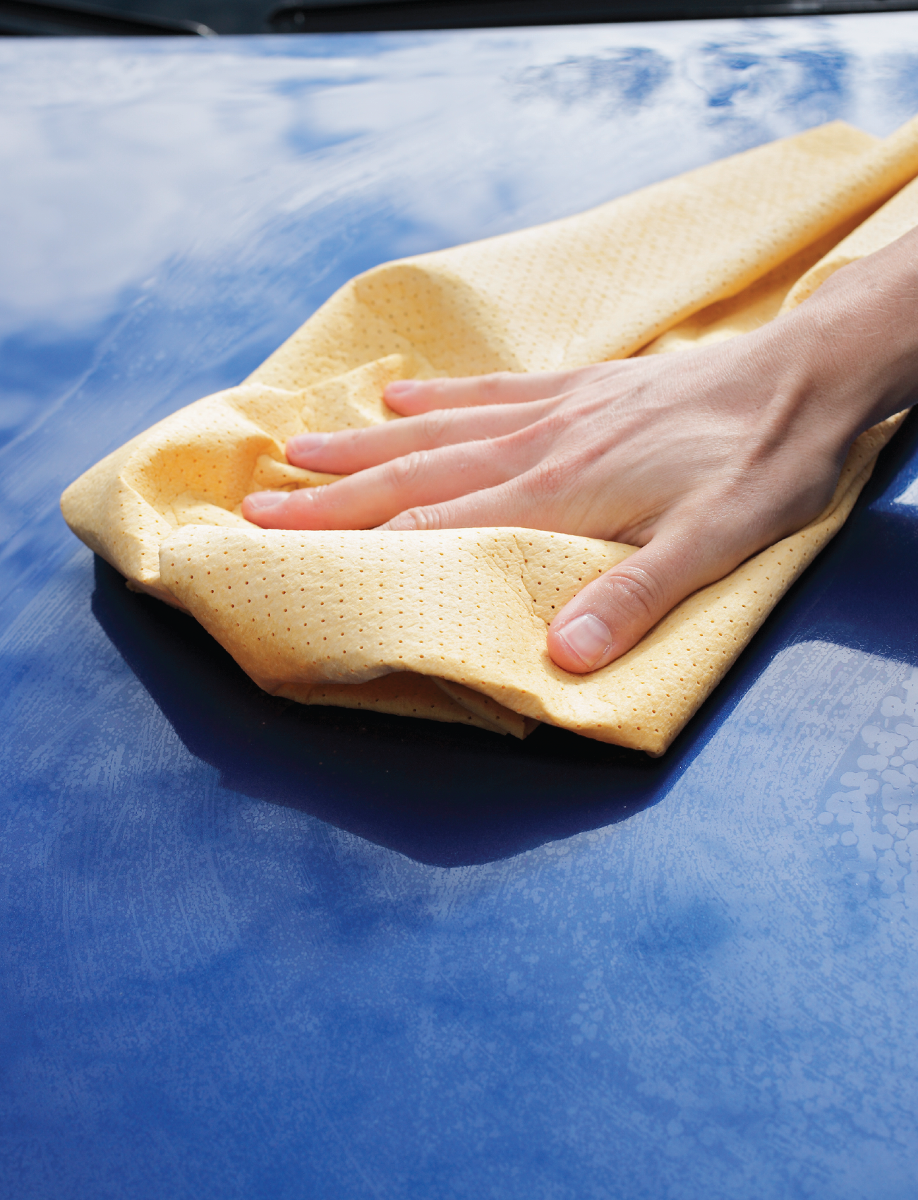A hand polishes a car's blue surface with the yellow Sunland-Protection CHAMOIS SYNTHETIC PVA 43 x 32cm, ensuring a streak-free shine while the car reflects the clear blue sky and clouds above.