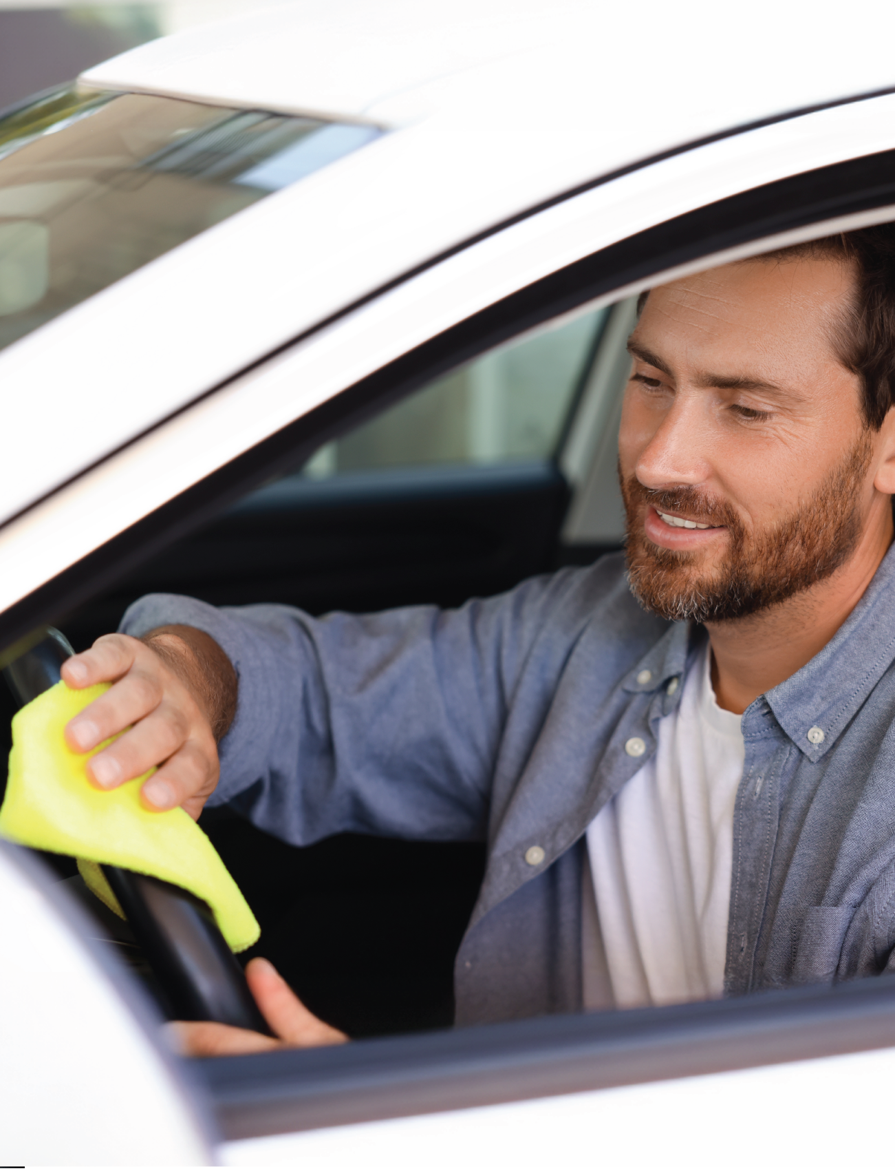 A man in a blue shirt sits inside a white car with the window open, smiling as he polishes the steering wheel with a yellow cloth. The blurry background complements the bright gleam of his car, thanks to Sunland-Protection's streak-free CHAMOIS SYNTHETIC PVA (43 x 32cm).