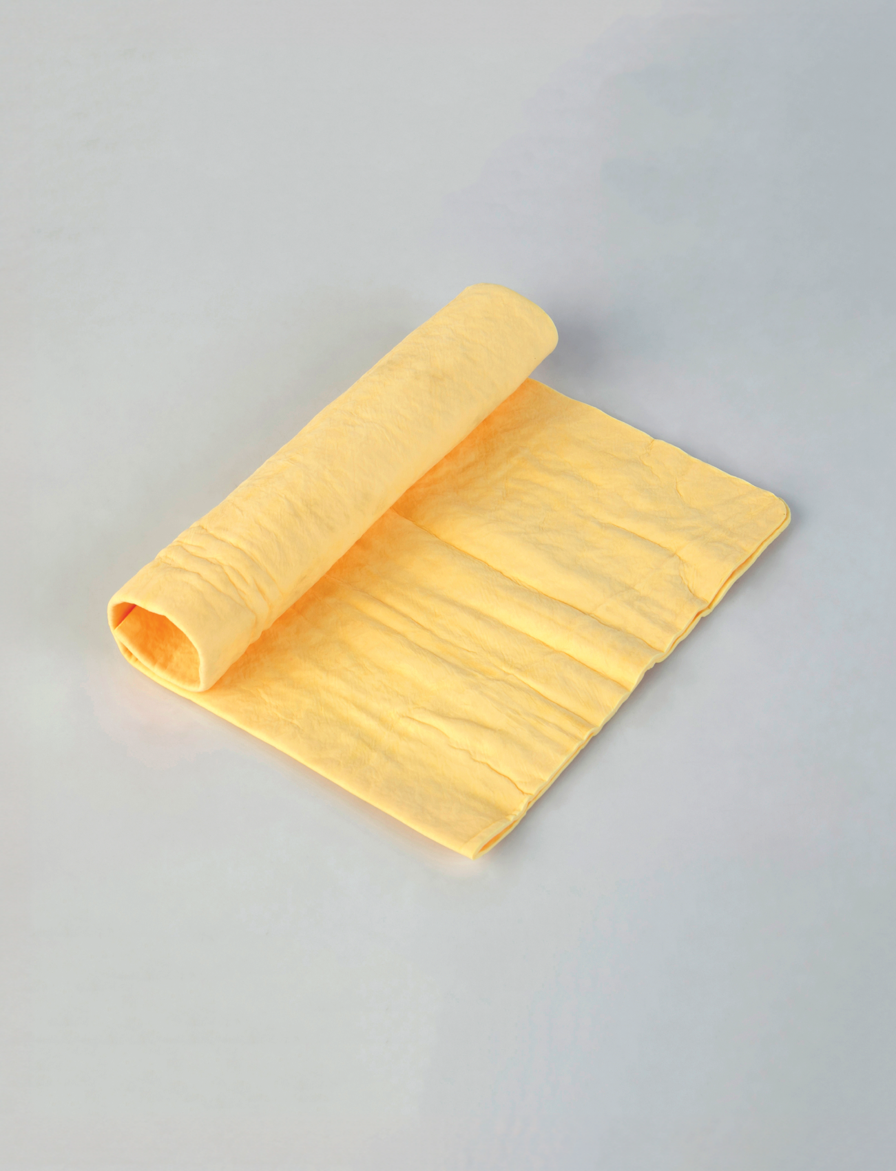 A CHAMOIS SYNTHETIC PVA from Sunland-Protection, measuring 43 x 32cm, lies neatly folded on a smooth light gray surface. Soft and textured, with one end rolled up, its streak-free design is ideal for detailing cars or cleaning delicate surfaces. Its yellow shade enhances its pristine touch.
