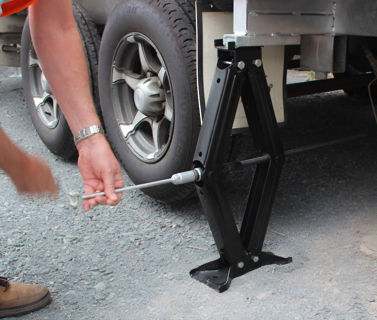 Using an Explore Caravan Jack, a person positions the jack under the vehicle near the rear wheel and uses a long metal rod to turn its screw mechanism. This certified jack, commonly used in caravan setups, slightly elevates the vehicle on a gravel surface.