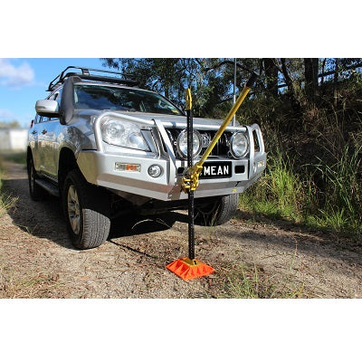 A silver off-road vehicle parked on a dirt path showcases recovery accessories, including a high lift jack securely mounted at its front bumper. The high lift jack, from the Mean Mother 4x4 brand and measuring 48 inches with an added bonus bag, is designed for stability and efficiency. Trees and grass appear in the background under a clear blue sky.