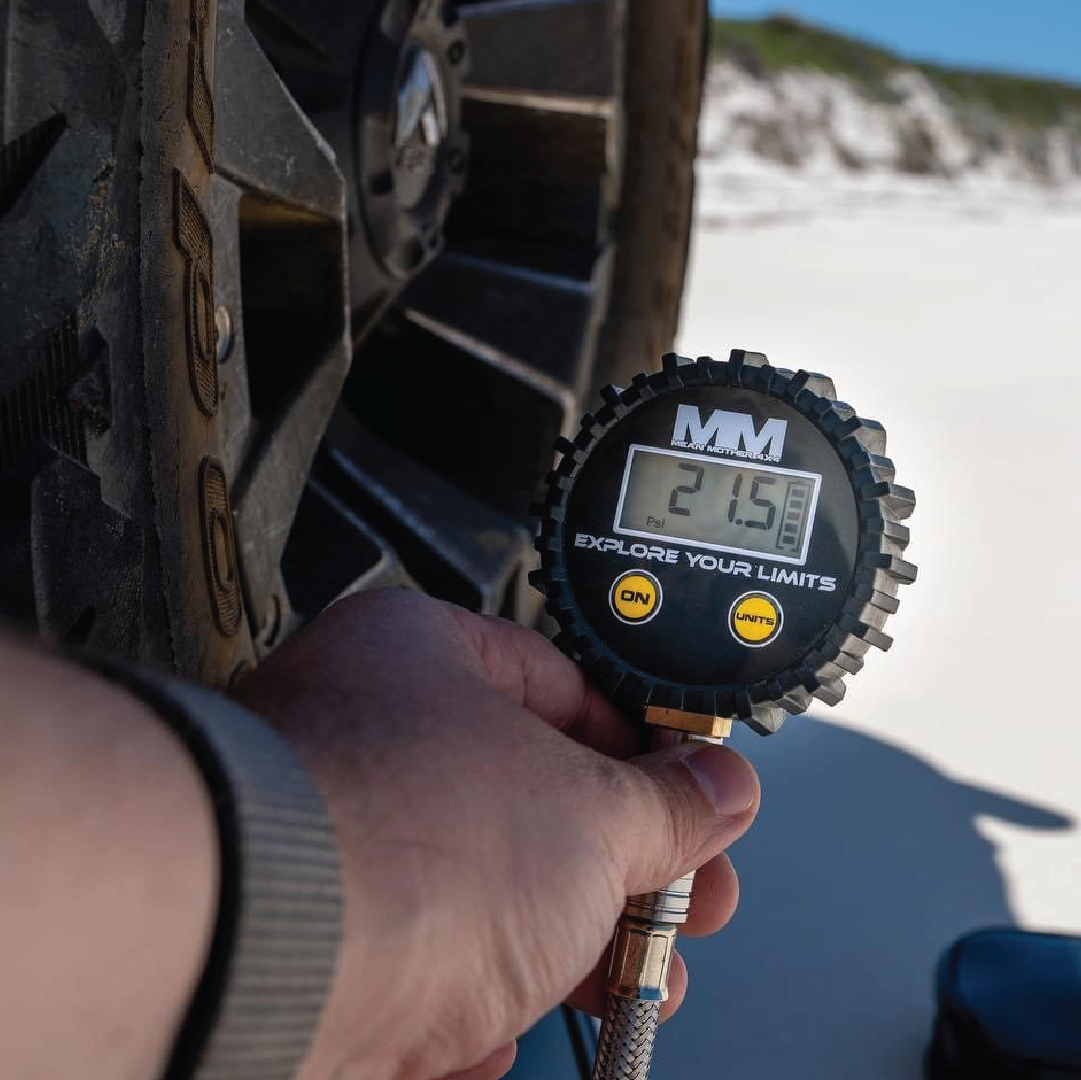 Someone is using a Mean Mother 4x4 Digital Tyre Deflator & Gauge Kit, displaying 21.5 PSI, to check a large vehicle tire's pressure against a backdrop of sandy landscapes and clear blue skies, hinting at beach driving adventures.