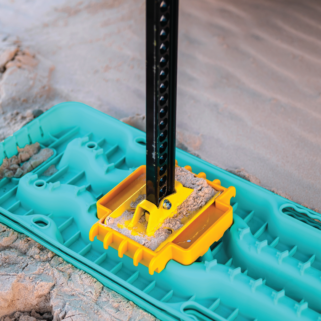 A metal post, reminiscent of the Mean Mother High Lift Jack 48", is anchored to a bright yellow mount on a textured turquoise base, set against a sandy surface. The vibrant colors stand out against the natural sand backdrop, evoking the image of recovery accessories ever ready for action.