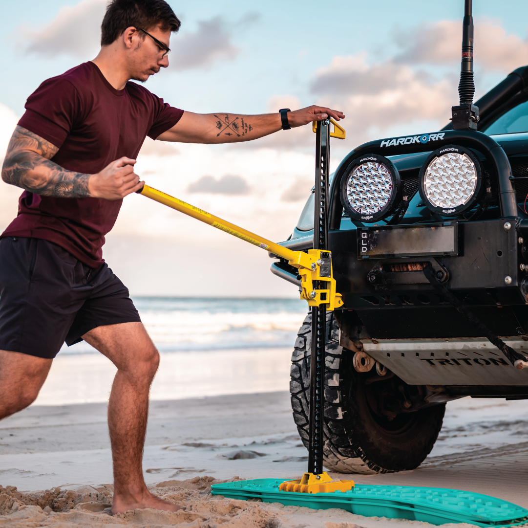 A casually dressed individual is utilizing the Mean Mother High Lift Jack 48", an essential recovery accessory, to elevate a 4x4 vehicle on a sandy beach. They are surrounded by the stunning scenery of the beach, with the ocean and sky serving as a beautiful backdrop.