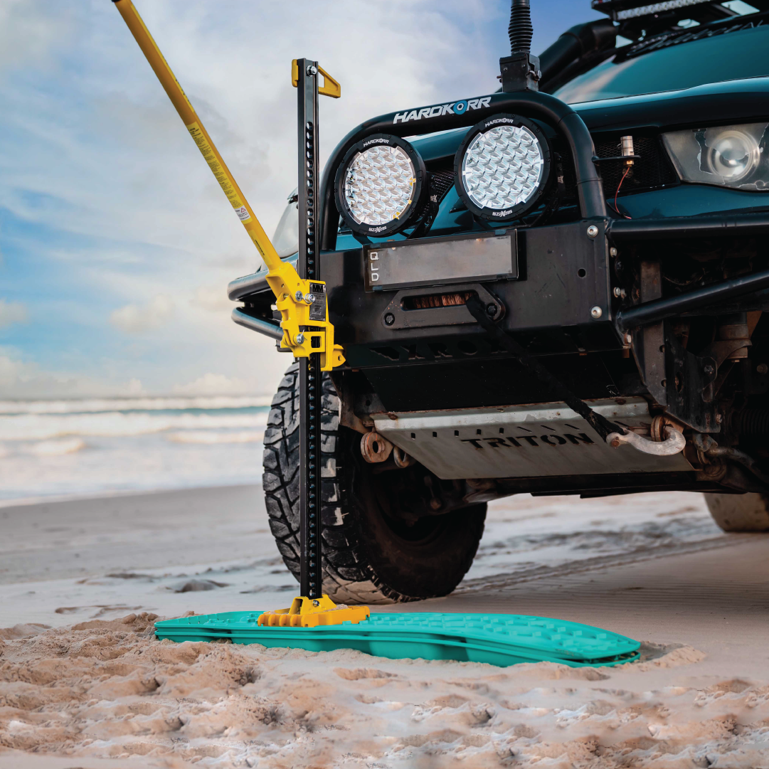On a sandy beach, an off-road vehicle utilizes the Mean Mother High Lift Jack 48", demonstrating its sturdy lifting capabilities for recovery. The jack is securely positioned on a bright turquoise recovery board, with the ocean and sky providing a stunning backdrop.