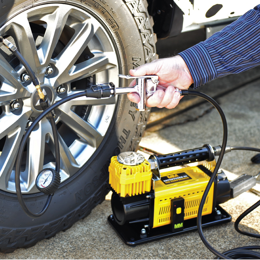 A person uses a portable Mean Mother 4x4 12V Air Compressor Bypass Controller in yellow to inflate a car tire. Placed on the ground, the compressor provides an accurate pressure reading through the gauge attached to the hose, allowing for precise valve adjustments. The details of the tire and vehicle's rim are clearly visible.