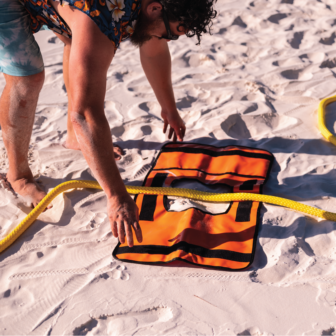 On a sandy beach, an individual clad in a floral shirt and barefoot is using a Mean Mother Recovery Dampener Blanket, distinguished by its high-visibility safety orange color and window feature. Nearby, a yellow hose rests on the sand amid various safety accessories.