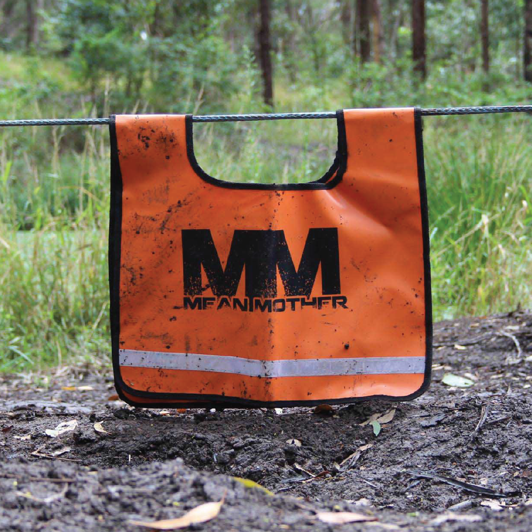 In a forest setting, an orange Mean Mother Recovery Dampener Blanket featuring the "Mean Mother" brand name hangs on a wire. This high-visibility safety accessory is dirty from outdoor use and has a reflective strip at the bottom, surrounded by trees and greenery as it serves its potential role as a winch cable dampener.