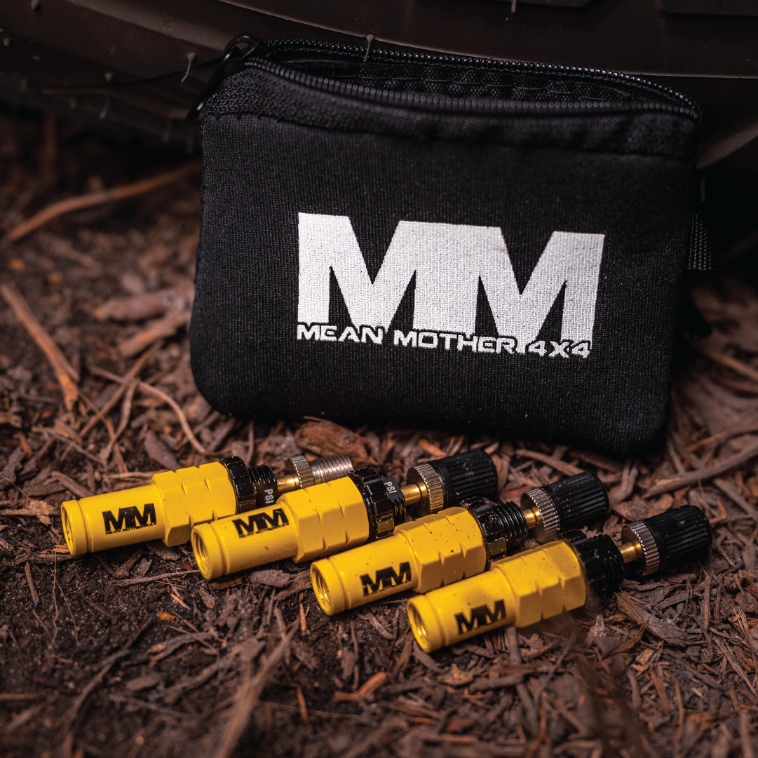 On the ground, ready for off-road escapades, are yellow tire deflators featuring black caps. A black zippered pouch, showcasing the "Mean Mother 4x4" label, takes center stage—ideal for any 4WD enthusiast. The surrounding twigs and dirt enhance the rugged allure of this outdoor setting.