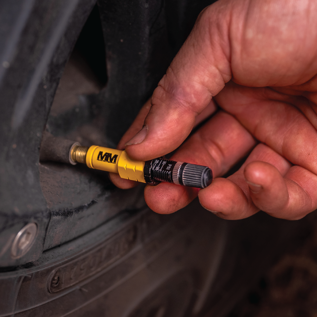 A close-up of a slightly dirty hand affixing a yellow and black air pump nozzle to a tire valve captures the essence of Mean Mother 4x4 Tyre Deflators. The partially visible off-road tire, marked for adventures, reveals the rugged texture of the rubber and wheel rim, hinting at upcoming 4WD excursions.