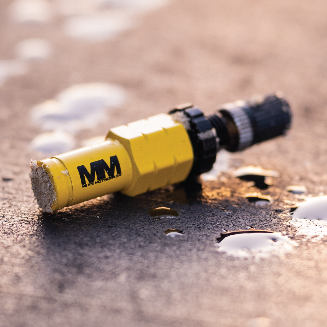 Close-up of a yellow cylindrical object with a black connector, reminiscent of the Tyre Deflators - 4 Pack by Mean Mother 4x4, often used in off-road adventures. The letters "MM" are printed on the yellow part, surrounded by water droplets on the wet surface. Warm and soft lighting enhances its rugged 4WD appeal.