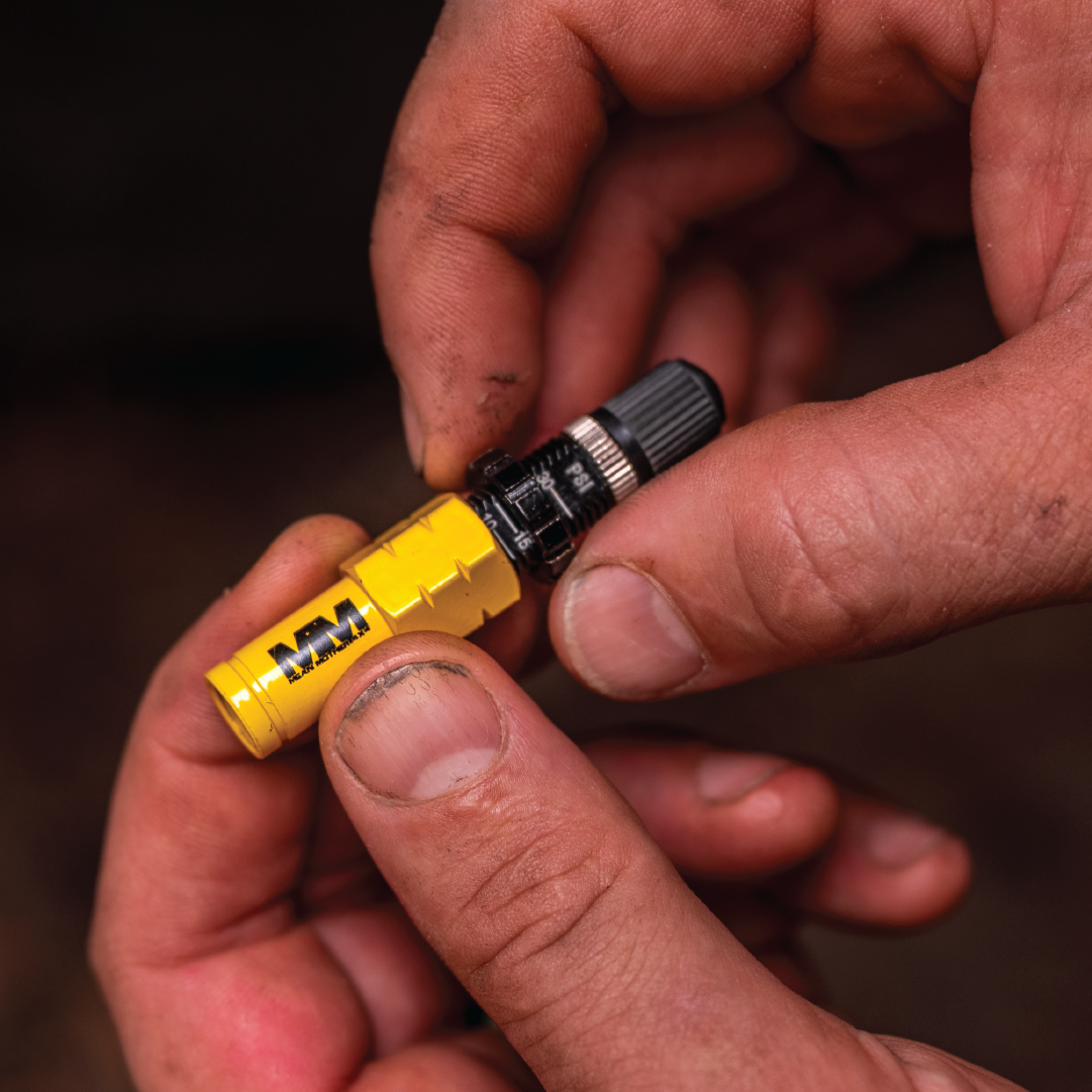 Close-up of a person's hands holding a Mean Mother 4x4 Tyre Deflator, featuring yellow and black accents with gears and a knob. The slightly dirty hands suggest a workshop or off-road setting, possibly working on 4WD equipment.