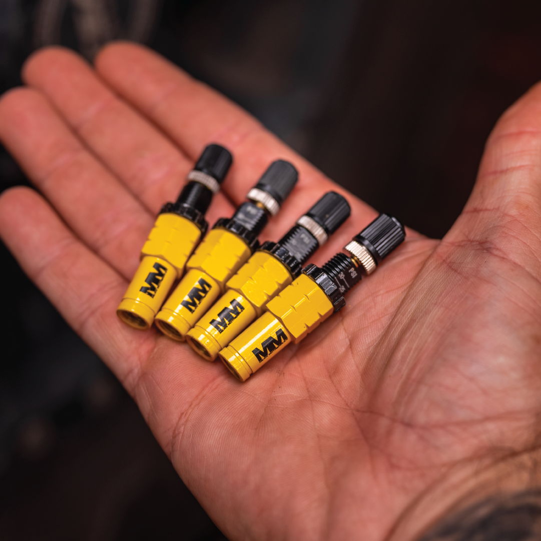 A firm hand grips four Tyre Deflators from the Mean Mother 4x4 brand, stacked parallel like rugged explorers conquering off-road terrains. Their compact design and sturdy metal construction are sharply in focus against a blurred backdrop.