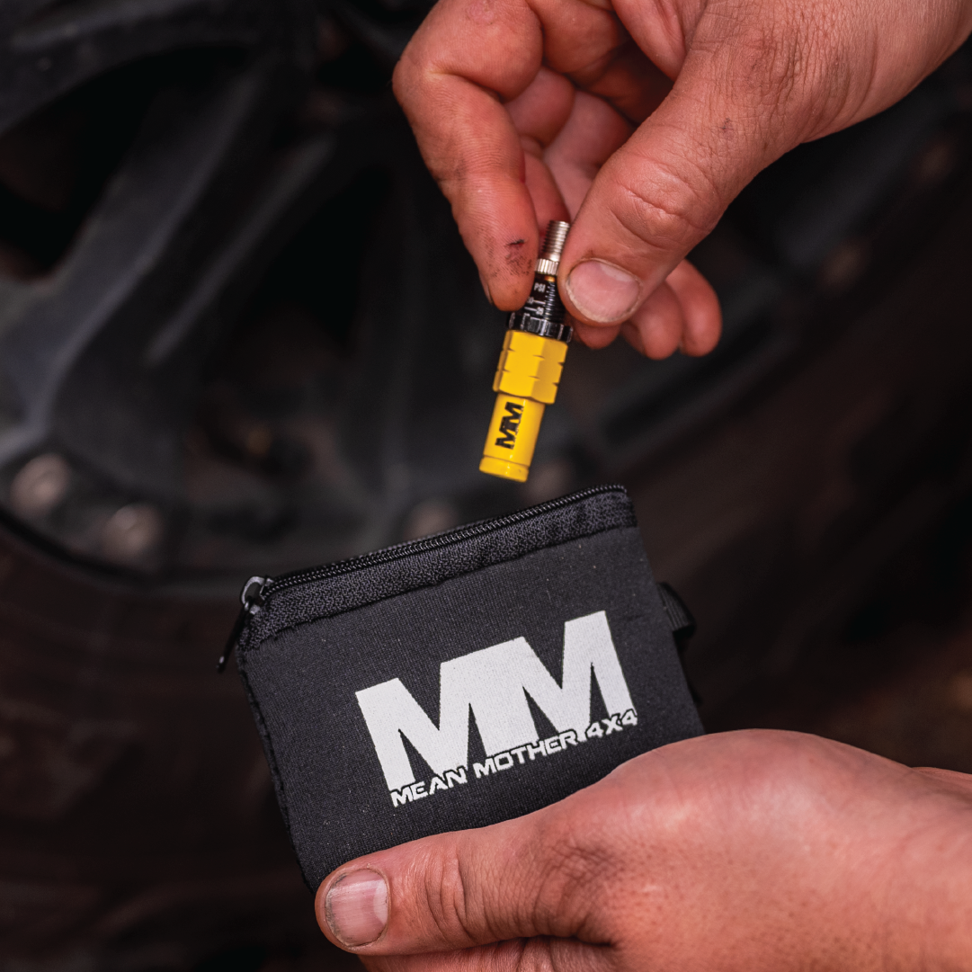 A person is holding a yellow deflator from the Tyre Deflators - 4 Pack by Mean Mother 4x4 near a tire, preparing to store it in a black pouch with "MEAN MOTHER 4X4" printed on it. Perfect for 4WD adventures, this tool allows off-road enthusiasts to efficiently manage their tyre pressure on the go.