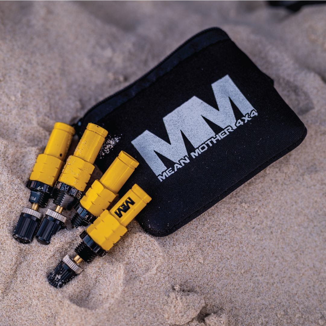 A set of Tyre Deflators - 4 Pack, designed by Mean Mother 4x4 for off-road enthusiasts, is half-buried in sand beside a black pouch displaying the "MM Mean Mother 4x4" logo. The deflators feature a striking yellow and black color scheme, ideal for 4WD adventures.