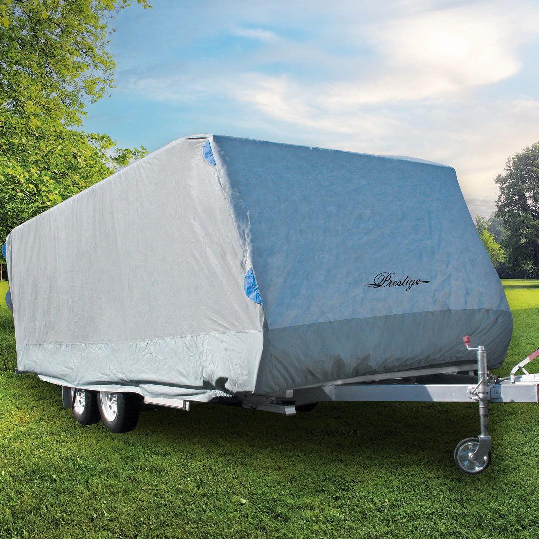 A large trailer is shielded by a gray, waterproof Prestige Caravan Cover 5.4m - 6.0m, designed with UV stabilization. It rests on green grass with trees and a partly cloudy sky in the background.