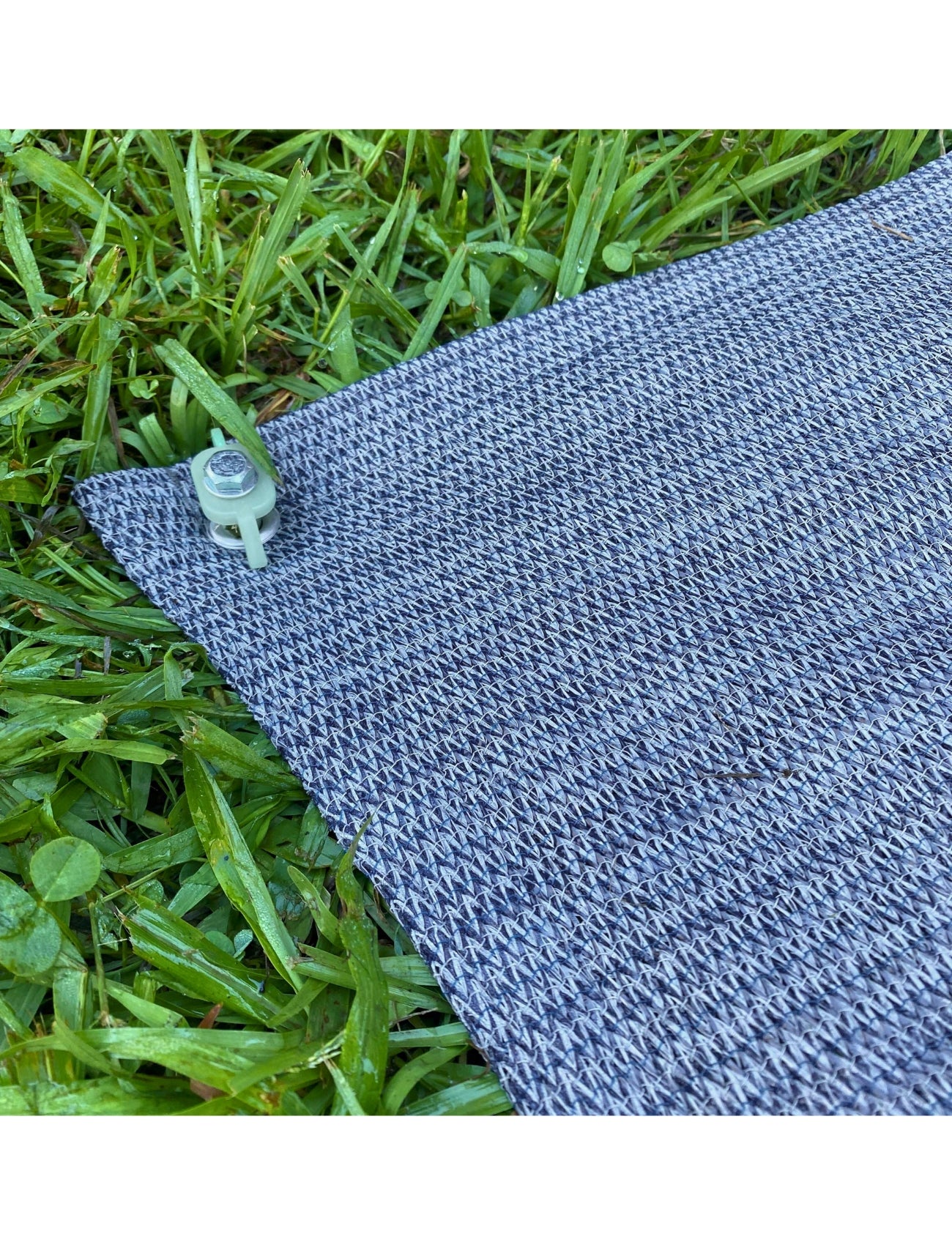 A close-up of a corner of the Sunland-Protection CARAVAN ANNEXE & FLOOR MATTING 2.5 x 3m, featuring a textured gray surface, secured with a small, round, transparent peg. The slip-resistant matting is laid on vibrant green grass with visible blades and a few dewdrops.