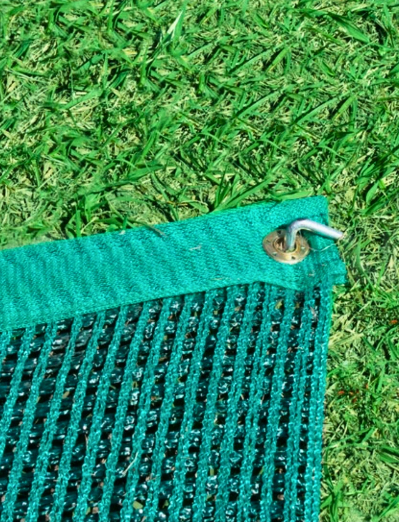 A Sunland-Protection ANNEX MATTING, measuring 2.5m x 5.0m in green and resembling polyethylene matting, is secured to the grass with a metal hook in the top corner. This matting features an open mesh pattern, and the surrounding grass is bright green and well-trimmed.