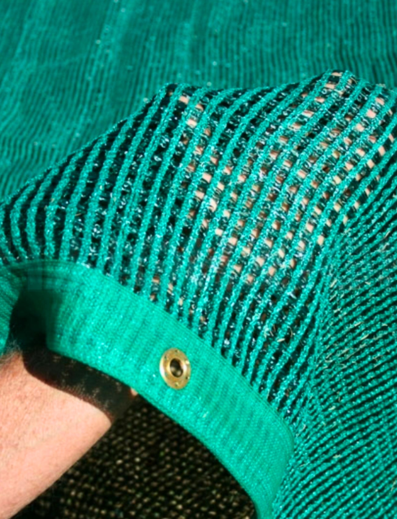 A detailed shot captures a person dressed in Sunland-Protection's ANNEX MATTING 2.5m x 7.0m GREEN, featuring black stripes that evoke the elegance of woven air. The material gleams with a metallic finish, highlighted by a rolled-up sleeve adorned with a brass eyelet. Surrounding them is more of the same textured polyethylene fabric, enhancing the scene's unity.