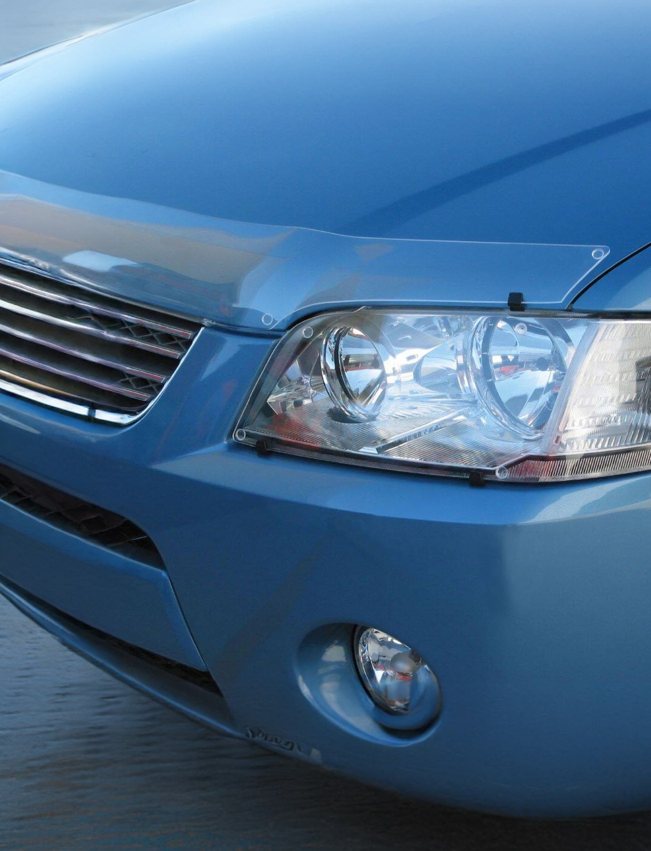 A close-up of the front corner of a light blue car showcases the headlight, grille, and part of the bumper. A Sunland Protective Plastics Bonnet Protector Clear for Ford Ranger PY (including Everest UB) 06/2022-On/> - F385B is elegantly attached, seamlessly integrating with the design in its custom-moulded, UV stable acrylic form.