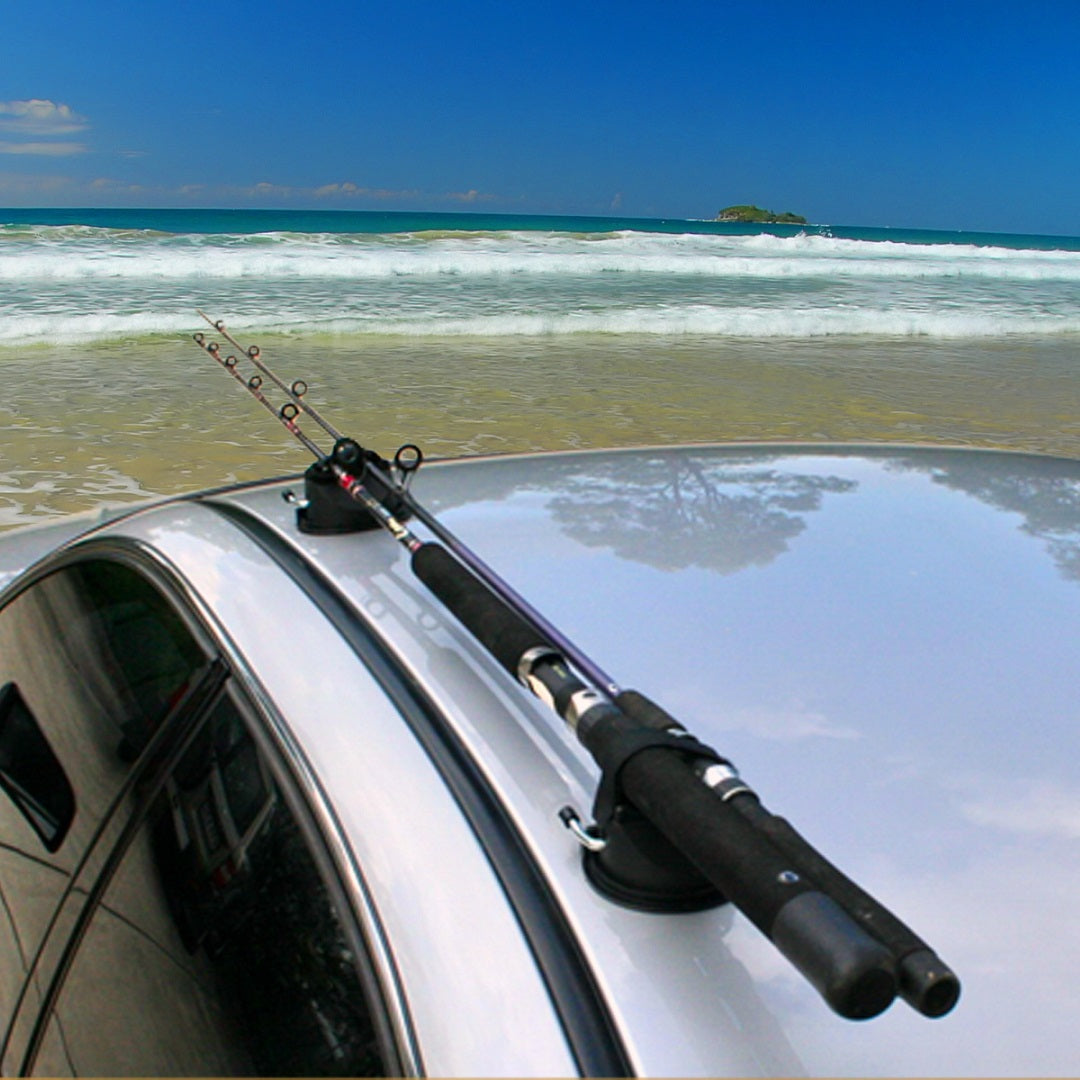 A pair of Cargo Mate Fishing Rod Holders securely fastens a fishing rod on top of a car. The car sits parked on a sandy beach, with gentle waves and a blue sky forming the picturesque backdrop. An island is visible in the distance.