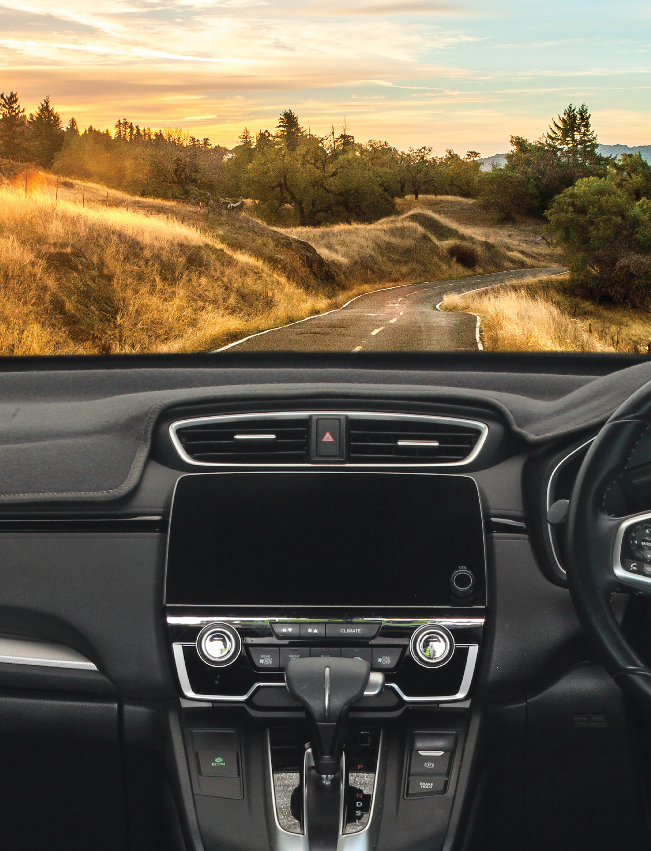 View from inside a car showing the dashboard and steering wheel, equipped with a Sunland-Protection Dash Mat Charcoal Suits Nissan Qashqai J12 10/2022-On/> With HUD - D88H06 to protect the vehicle dash and reduce glare. The winding rural road ahead is surrounded by dry grass and trees, leading towards a distant horizon with a colorful sunset sky.