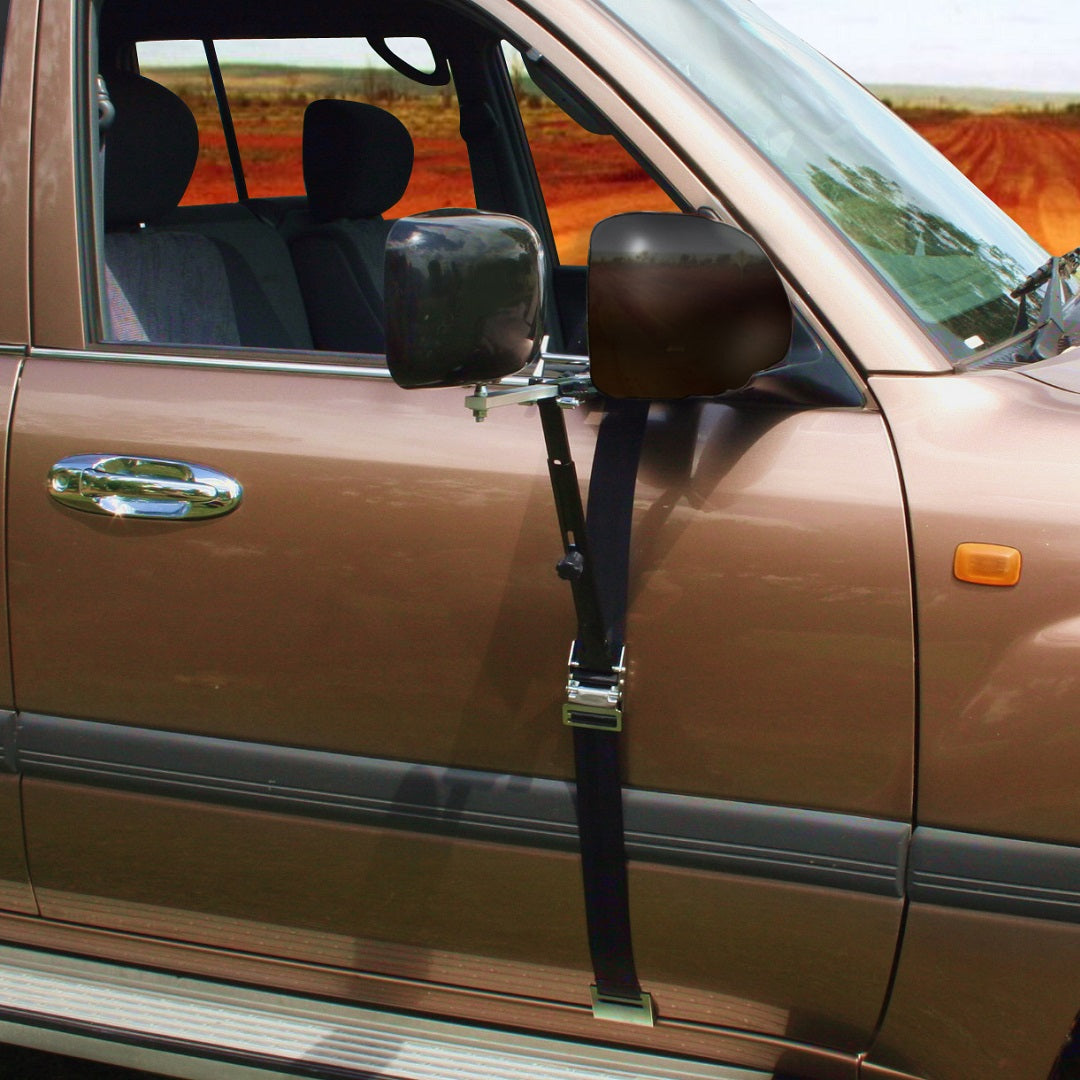 A tan-colored car with a seat belt fastened to the side mirror, tied around the front door. The car is parked on a dirt road with an open field in the background. The seat belt, coupled with a Drive Towing Mirror from Drive for superior rear vision, appears to serve as an improvised security measure.