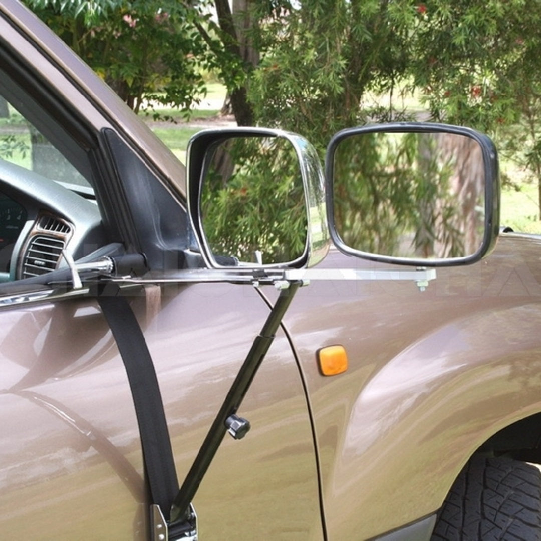 Close-up of a car's side mirror featuring the Drive Towing Mirror by Drive, designed for superior rear vision and secured with adjustable straps and brackets. This heavy-duty towing mirror includes an adjustable ratchet mechanism for easy positioning. The car is parked outdoors near greenery.