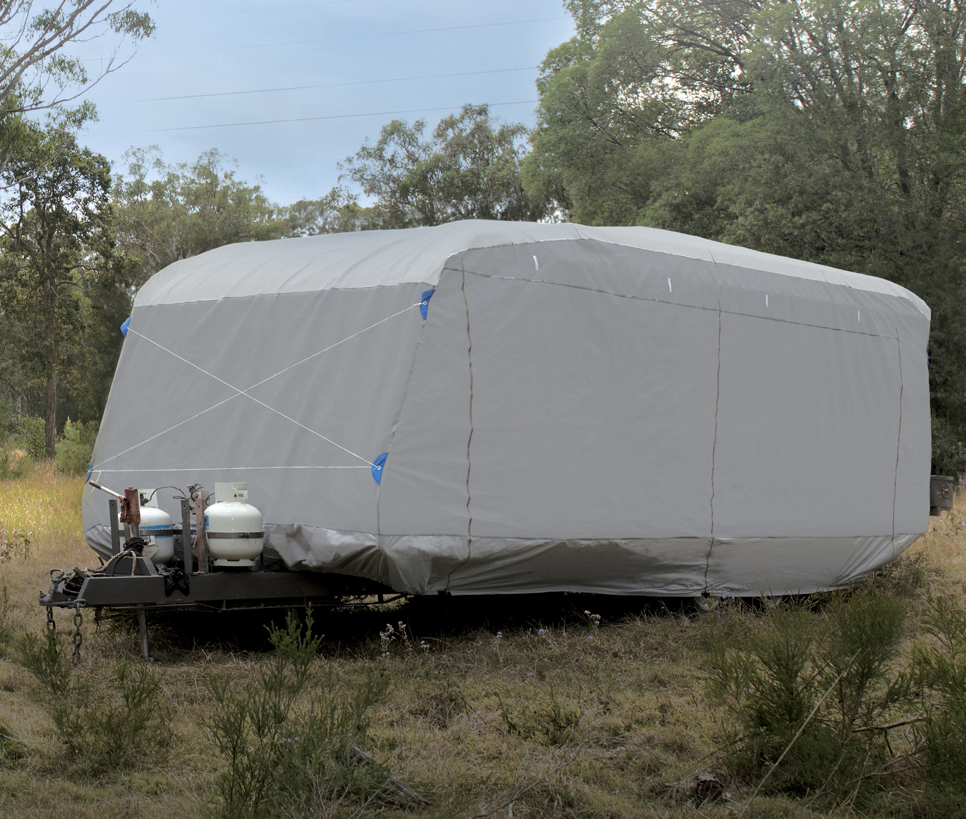 A large covered trailer sits in a grassy, wooded area. The trailer, wrapped in an Explore Caravan Cover 6.0m - 6.6m with reinforced stitching and a water-resistant top panel, is secured with ropes. Two propane tanks are mounted on the front of the trailer. Dense trees and foliage are visible in the background under a cloudy sky.