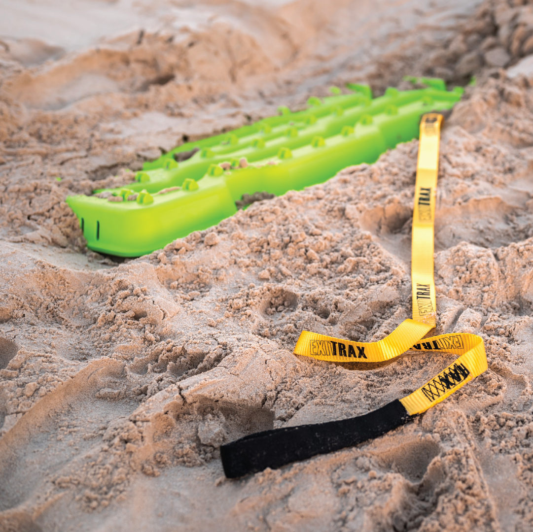 An Exitrax recovery traction board with a reinforced handle is partially buried in sandy terrain. Leading away from the brightly colored board is an Exitrax yellow and black recovery leash, partially extended across the sand. The scene appears to be set on a beach or desert-like environment.