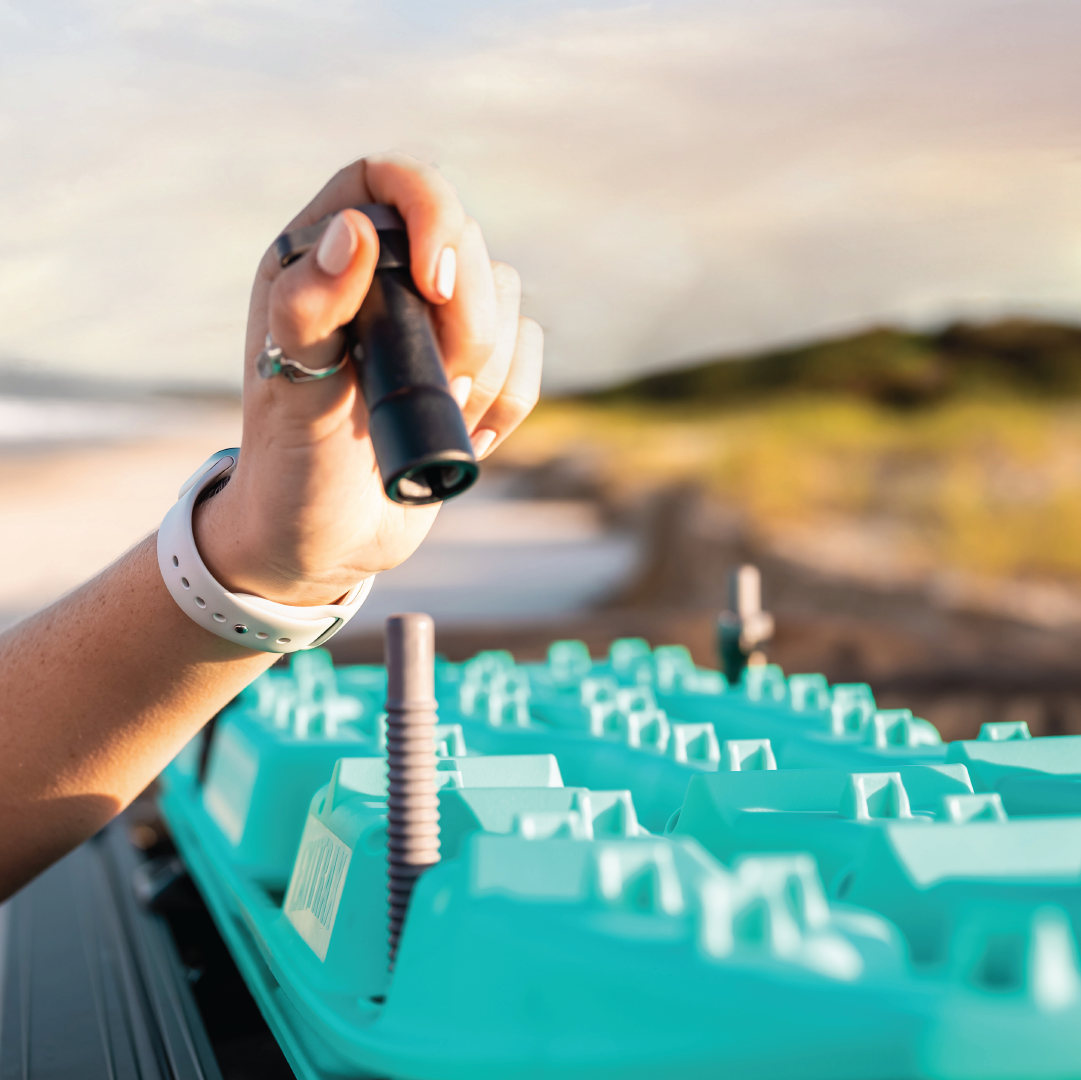 Close-up of a person's hand holding a flashlight over an open battery pack outdoors. The person is wearing a white wristband and a silver ring. The background shows blurred greenery, a body of water under a light blue sky, and Exitrax recovery boards mounted using the Exitrax Recovery Board Mounting Kit in the distance for quick emergency use.