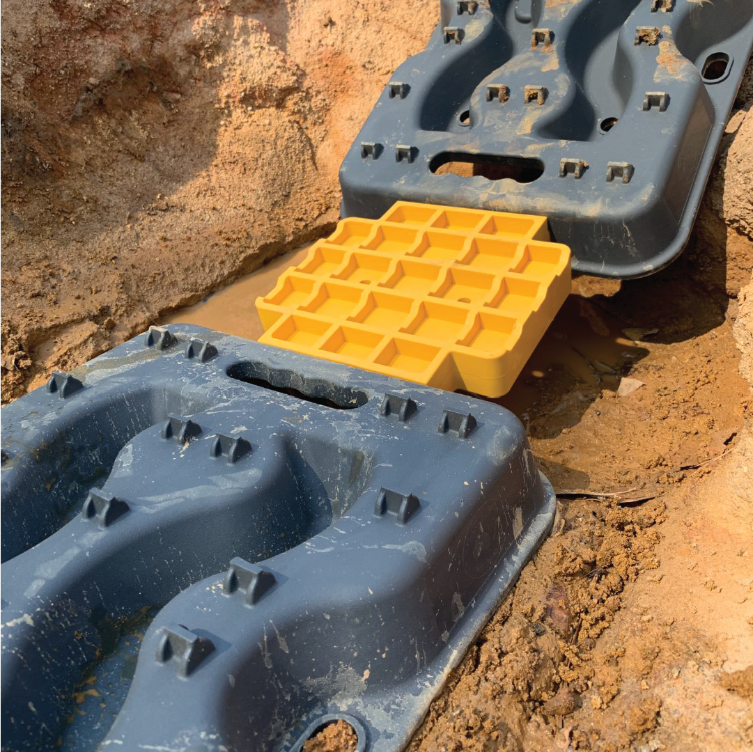 Close-up of blue and yellow Exitrax Recovery Board Link Plates / Jack Bases (Exilink) used to stabilize and level a surface in a construction site, placed on muddy ground. The blue pieces have handle cutouts and raised grips, while the yellow piece features a grid pattern.