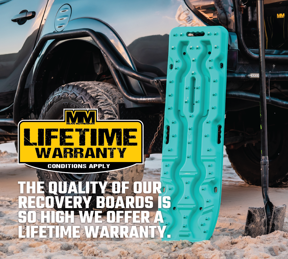 An Aqua Marine Exitrax Recovery Board Ultimate 1150 stands upright in the sand next to a shovel and a black off-road vehicle. A large yellow and black sign reads, "MM Original Lifetime Warranty Conditions Apply." Text underneath states, "The quality of our Exitrax recovery boards is so high we offer a lifetime warranty.