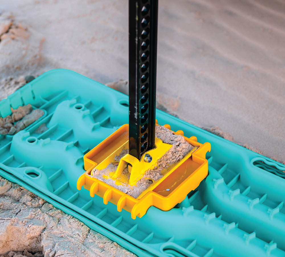 A close-up of a yellow jack on the Exitrax Recovery Board Ultimate 1150 - Aqua Marine, part of a pair and bundled with Recovery Board Mounts, featuring a metal pole resting on the turquoise mat in the sand. The mat is designed with grooves and textured surfaces for grip, accentuated by its surrounding sandy landscape.