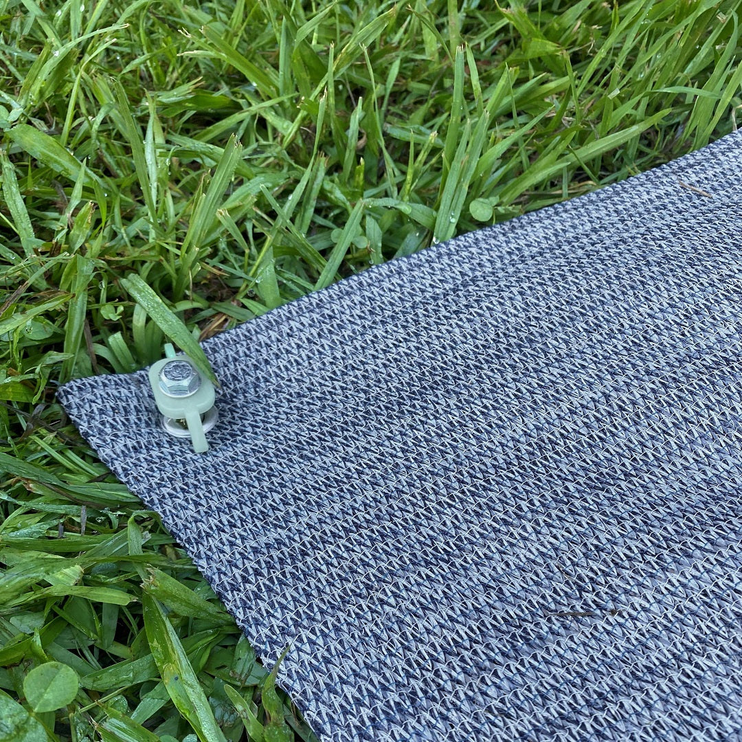 A close-up of the black and white woven Explore Caravan & Floor Matting 2.5 x 3m, featuring a mesh design, pinned to green grass with a plastic clip.