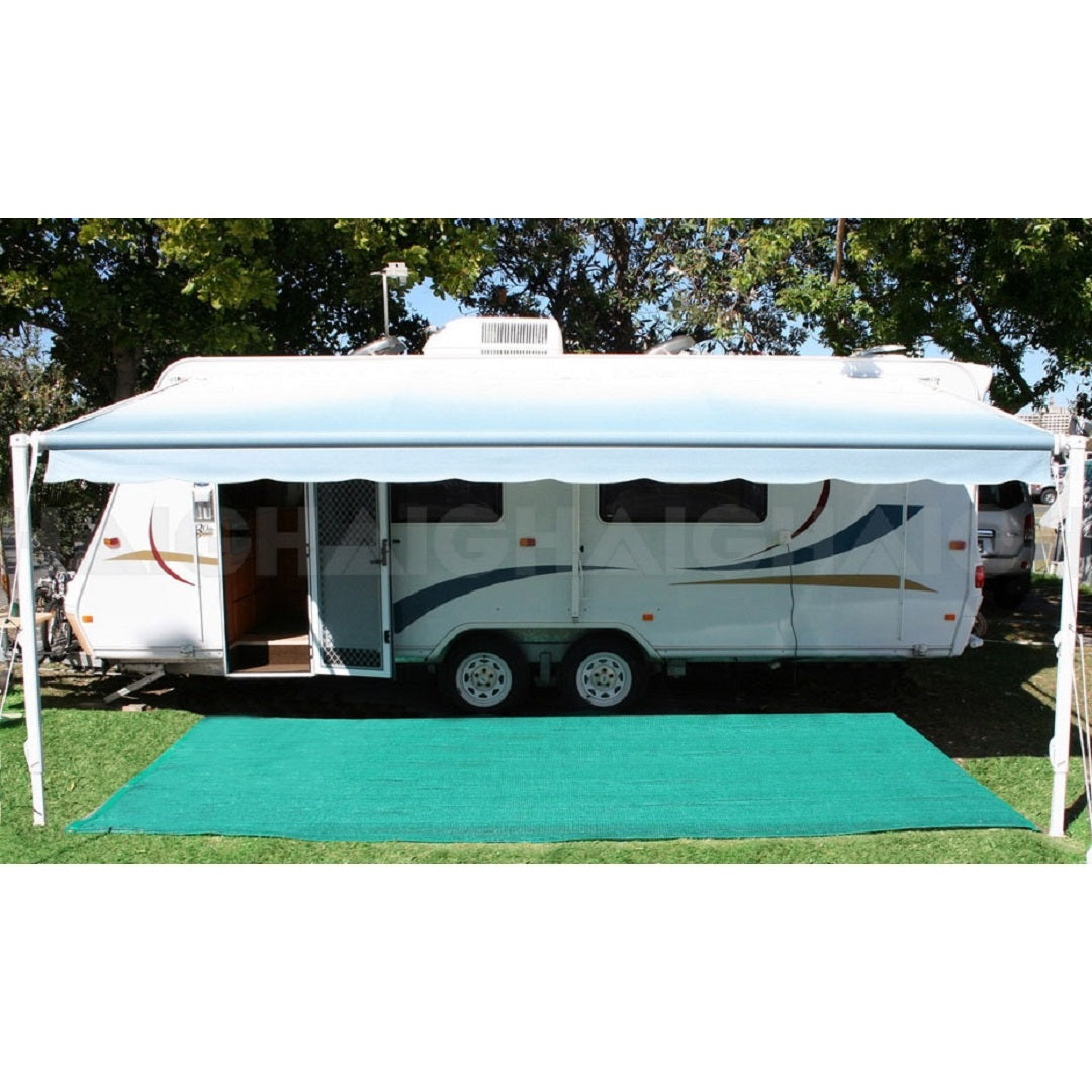 A white travel trailer with blue and orange accents is parked on grass, featuring a large blue awning extended for shade. Spread out in front of the caravan, underneath the awning, is the Explore Air Weave Annex Matting 2.5 x 5m by Explore. Trees are visible in the background.