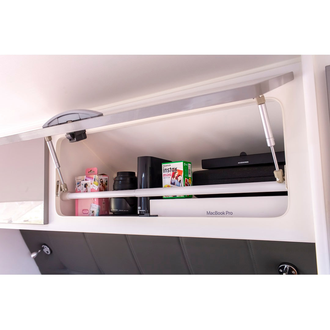 An open overhead cabinet reveals a well-organized collection: camera lens, "instax mini" film box, a MacBook Pro case, and a pink box neatly lined up like Explore Caravan & RV Fridge Bars from 48-80cm by Explore, ensuring everything stays in place on the white shelf.