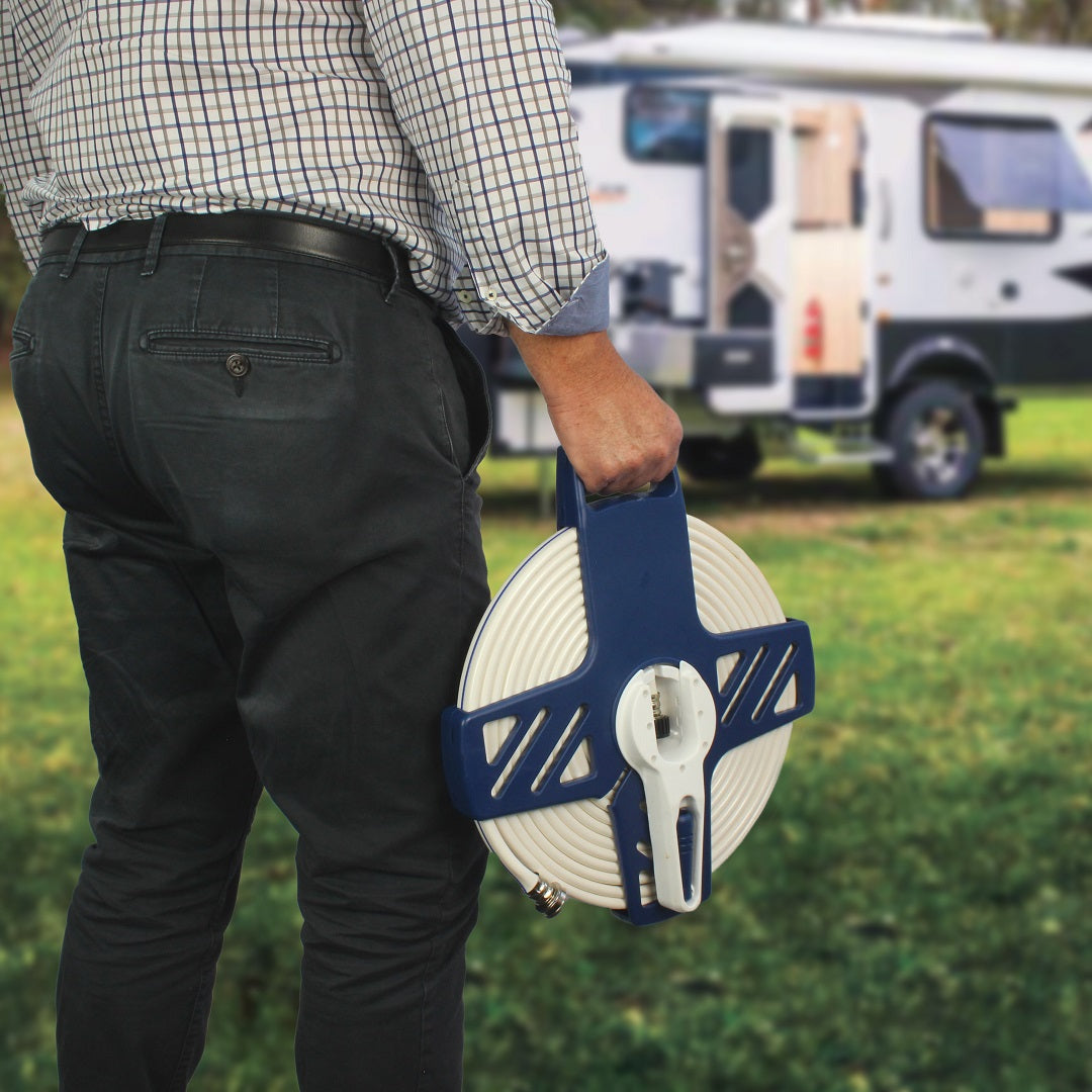 A person standing outdoors is holding an Explore Flat Hose With Reel - 9m in their hand. A parked RV is visible in the background on a grassy area. The person is dressed in dark pants and a checkered shirt.