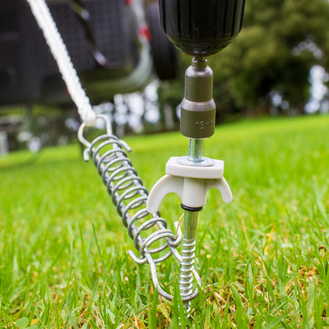 A close-up of an outdoor scene depicting an Explore power drill driving an Explore Hard Surface 8 Pce Metal Screw Peg into the hard ground. The metal screw peg, connected to a metal spring and chain, secures an unseen object. The grassy ground and blurred background with trees suggest a park or backyard.