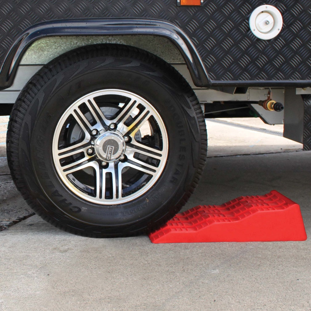 A close-up view of a vehicle tire resting on a black alloy wheel parked on a concrete surface. The tire is secured by the Explore Caravan Levelling Ramp - Australian Made, featuring a bright red, UV-resistant, textured surface placed behind the wheel to prevent rolling. Part of the vehicle's underside and body is visible.