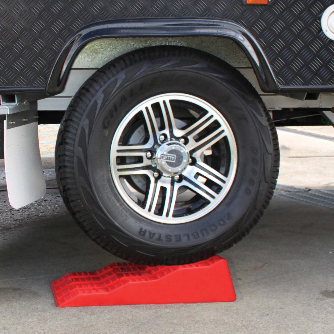 A vehicle wheel rests on the vividly colored Explore Caravan Levelling Ramp - Australian Made. This wheel belongs to a larger vehicle, probably a trailer or camper, that is outfitted with UV-resistant levellers by Explore. The background displays a paved surface along with sections of the vehicle’s wheel well and fender.