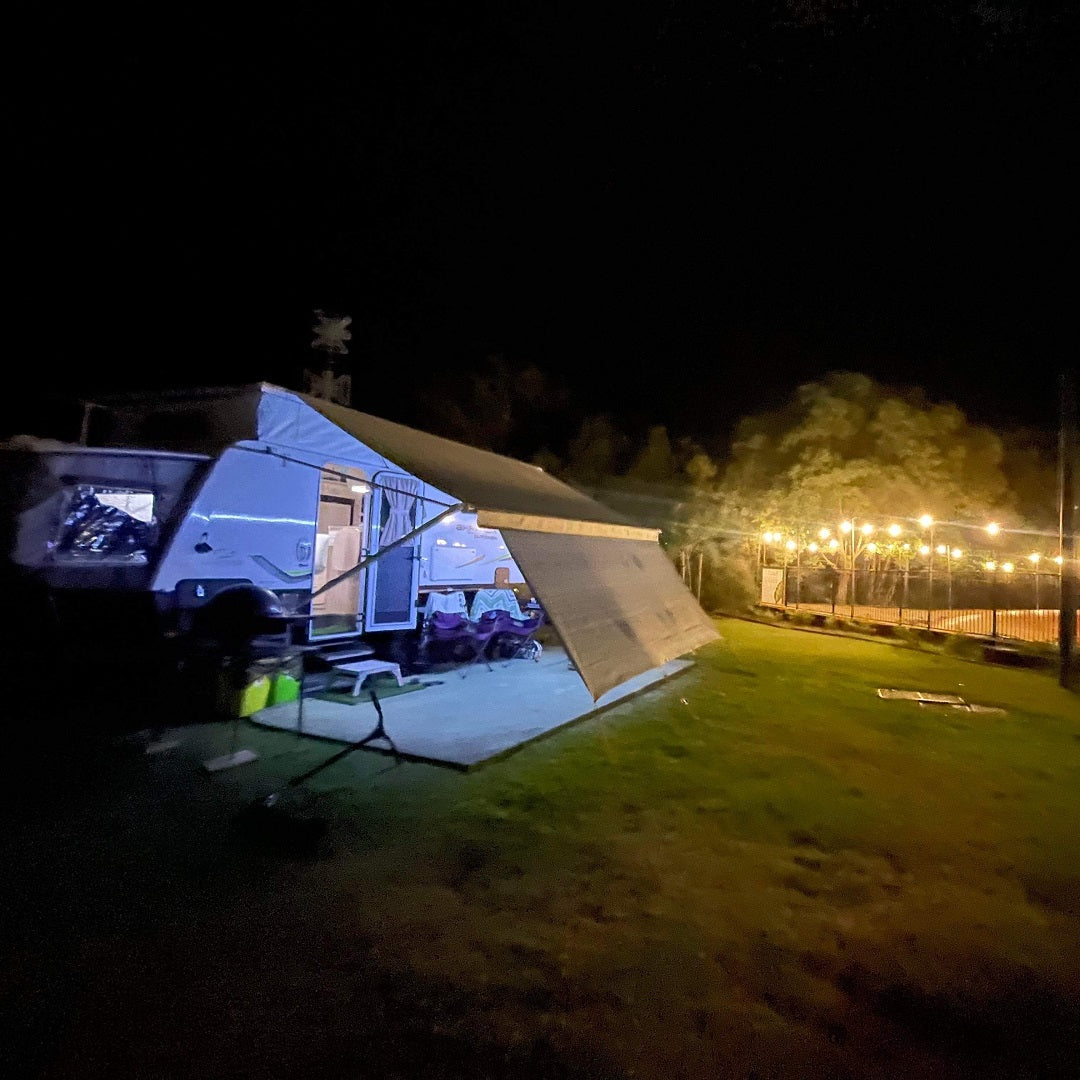 An Explore Caravan Privacy Screen 1.8m X 3.4m is set up to create a cozy nook among the trees as the camping trailer is extended at night with its awning out. The area is dimly lit, but string lights hanging nearby provide additional illumination to the grassy site, which is scattered with various camping gear.